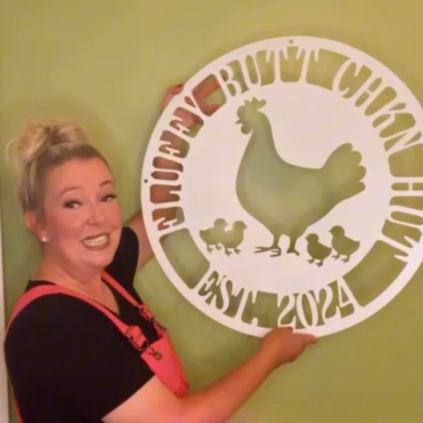 Woman holding a decorative metal sign with a rooster design against a green wall.