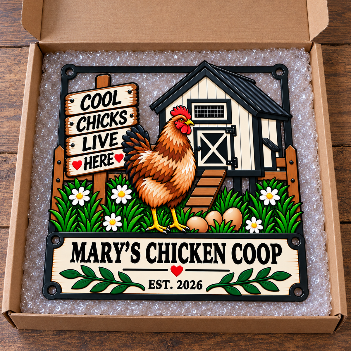 A rectangular decorative sign with a colorful chicken coop illustration and the text 'MARY'S CHICKEN COOP' and 'EST. 2026' sits inside an open cardboard box filled with bubble wrap on a wooden surface.