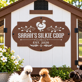A decorative brown and white wooden sign reading “SARAH'S SILKIE COOP” and “EST. 2026” hangs on the front of a white chicken coop.