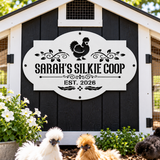 A decorative white sign with black lettering reading “SARAH’S SILKIE COOP” and “EST. 2026,” mounted on a black-and-white coop exterior.