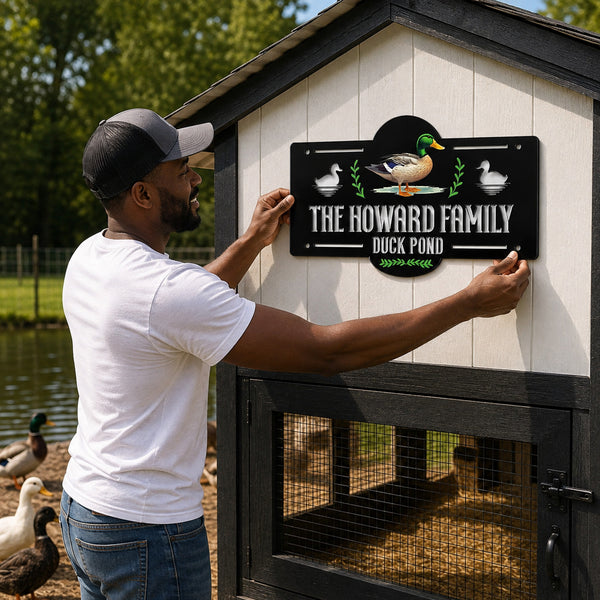 A man in a white t-shirt and jeans is installing a custom black metal sign on the wall of a small duck coop. The sign features a cartoon duck and the text 'THE HOWARD FAMILY DUCK POND'.