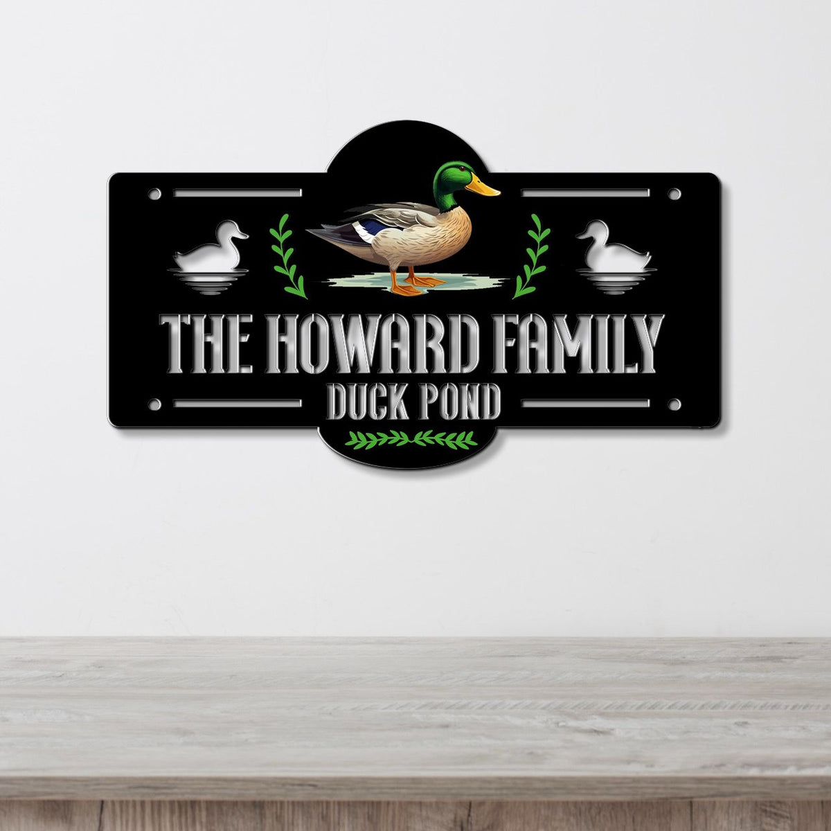 A decorative metal sign mounted on a light gray wall above a wooden surface. The sign is black with white text reading 'THE HOWARD FAMILY DUCK POND,' a mallard duck illustration in the center, white duck silhouettes on both sides, and green leafy accents.