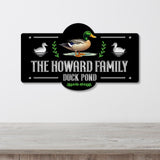 A decorative metal sign mounted on a light gray wall above a wooden surface. The sign is black with white text reading 'THE HOWARD FAMILY DUCK POND,' a mallard duck illustration in the center, white duck silhouettes on both sides, and green leafy accents.