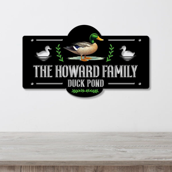 A decorative metal sign mounted on a light gray wall above a wooden surface. The sign is black with white text reading 'THE HOWARD FAMILY DUCK POND,' a mallard duck illustration in the center, white duck silhouettes on both sides, and green leafy accents.