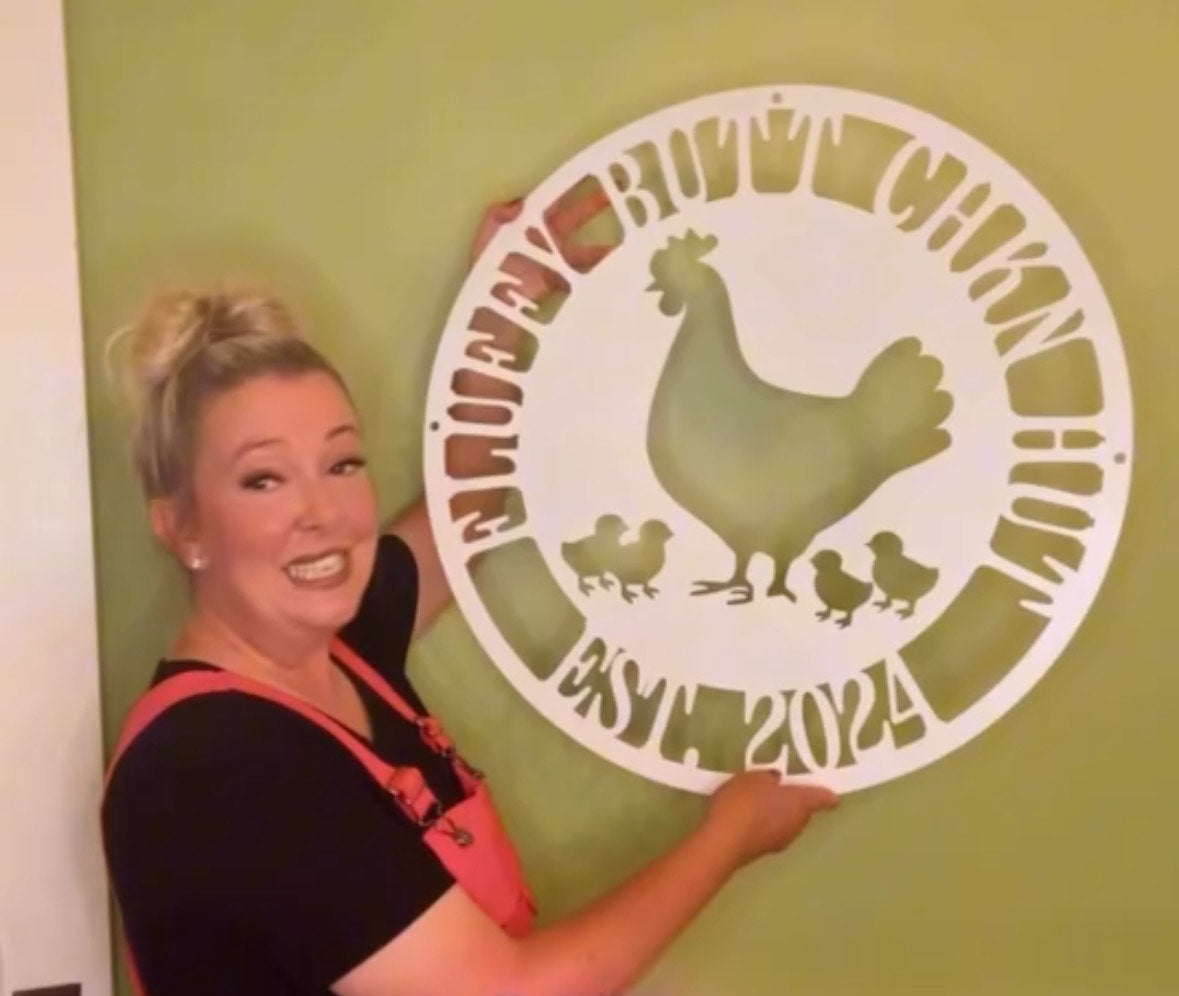 Woman holding a decorative metal sign with a rooster design against a green wall.