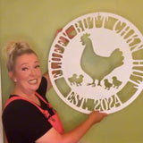 Woman holding a decorative metal sign with a rooster design against a green wall.