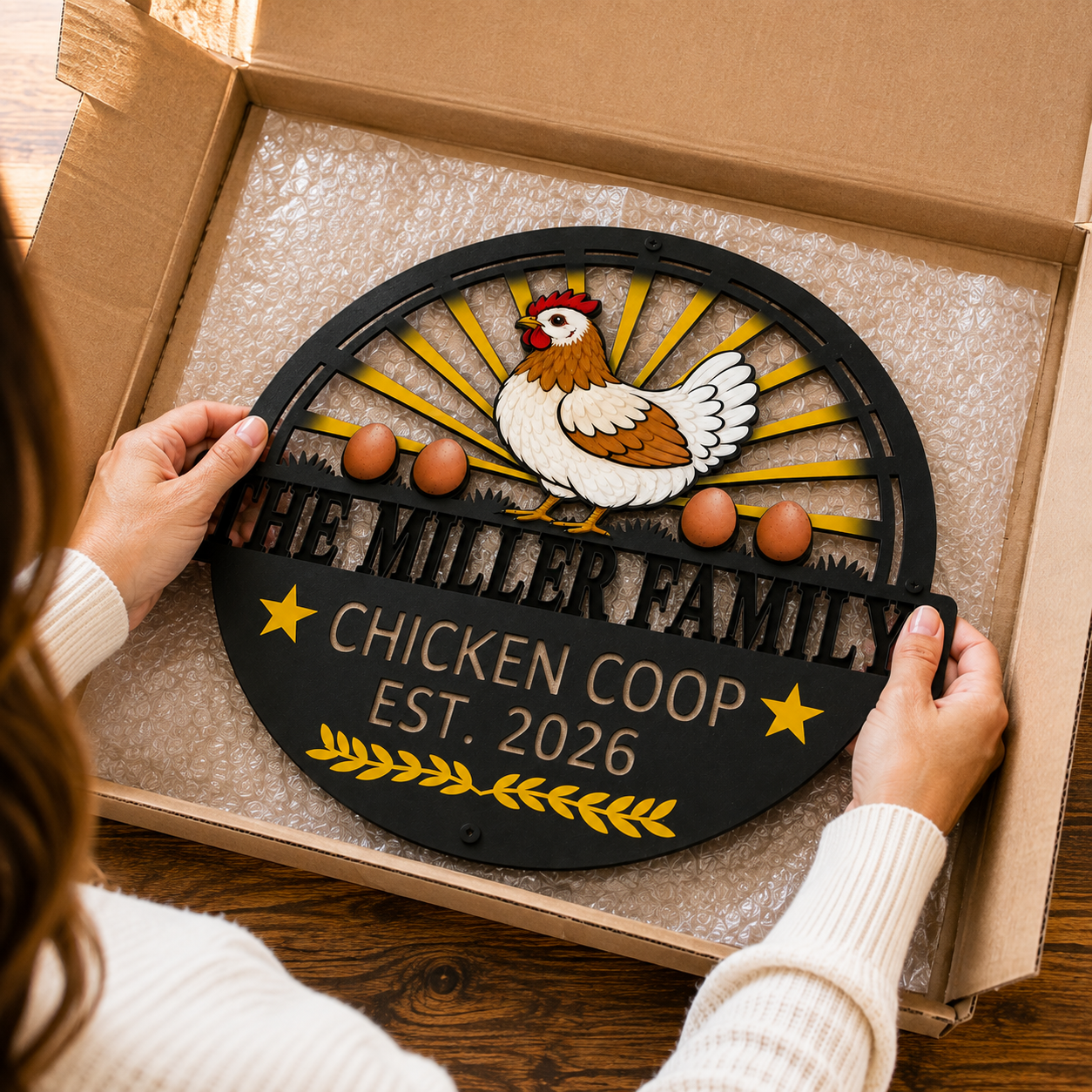 A person holds a round black metal sign inside a cardboard box. The sign features a white and brown chicken, yellow sunburst rays, three brown eggs, and the text 'THE MILLER FAMILY CHICKEN COOP EST.