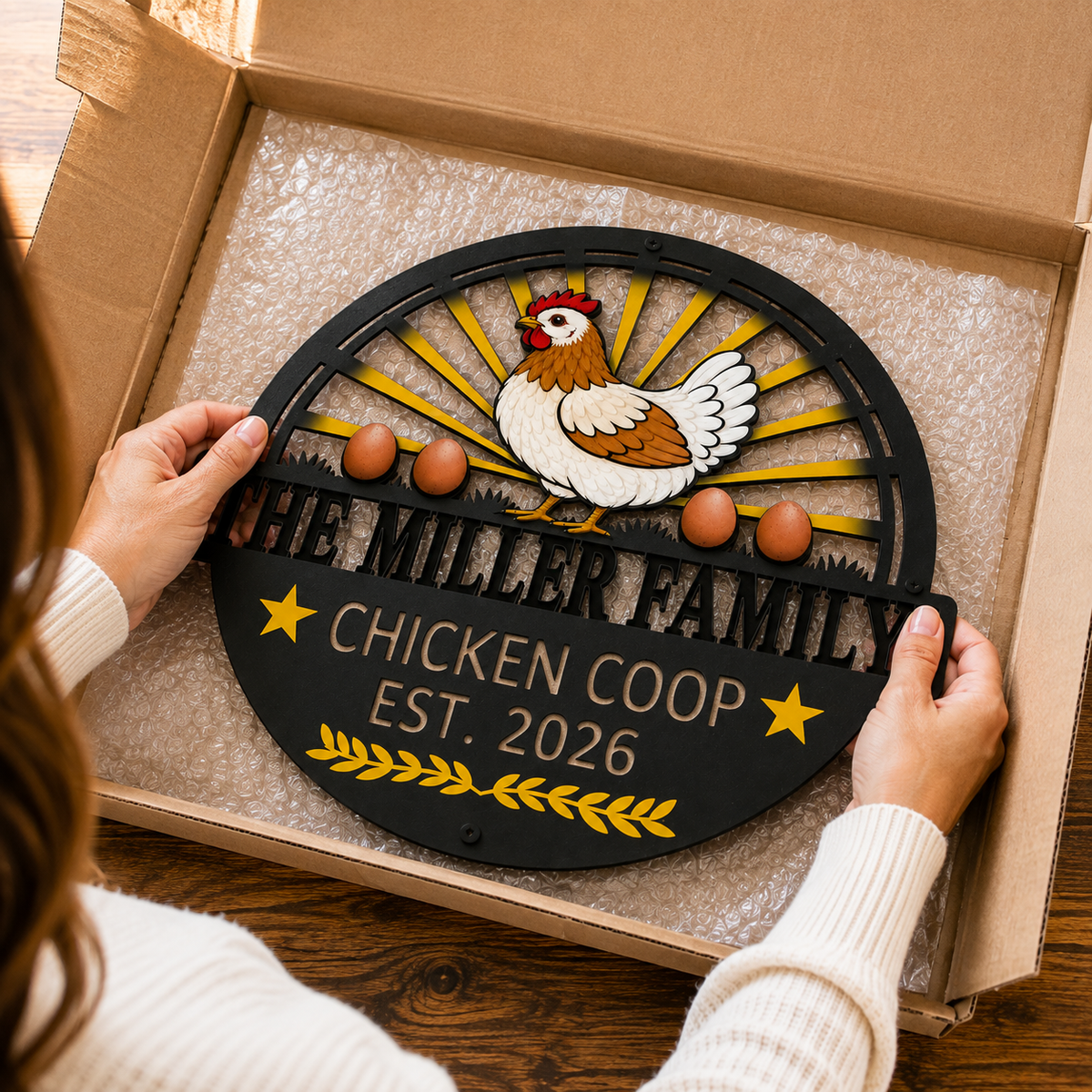 A person holds a round black metal sign inside a cardboard box. The sign features a white and brown chicken, yellow sunburst rays, three brown eggs, and the text 'THE MILLER FAMILY CHICKEN COOP EST.