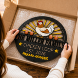 A person holds a round black metal sign inside a cardboard box. The sign features a white and brown chicken, yellow sunburst rays, three brown eggs, and the text 'THE MILLER FAMILY CHICKEN COOP EST.
