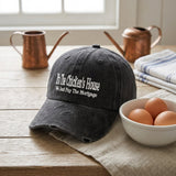 A dark gray distressed baseball cap with white embroidered text reading 'It's The Chicken's House We Just Pay The Mortgage' sits on a rustic wooden table styled with copper pitchers, a bowl of eggs, and a striped napkin.