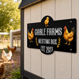 A decorative black metal sign for a chicken nesting box mounted on a white wooden coop. The sign features the text 'GABLE FARMS NESTING BOX EST 2023' in white, flanked by illustrations of two golden-brown chickens and small yellow chicks, with small gold leaf accents.