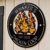 A circular black metal sign mounted on a beige wood-paneled wall, featuring cartoon chickens and the text 'JENNIFER'S CHICKEN COOP' and 'EST. 2026'.