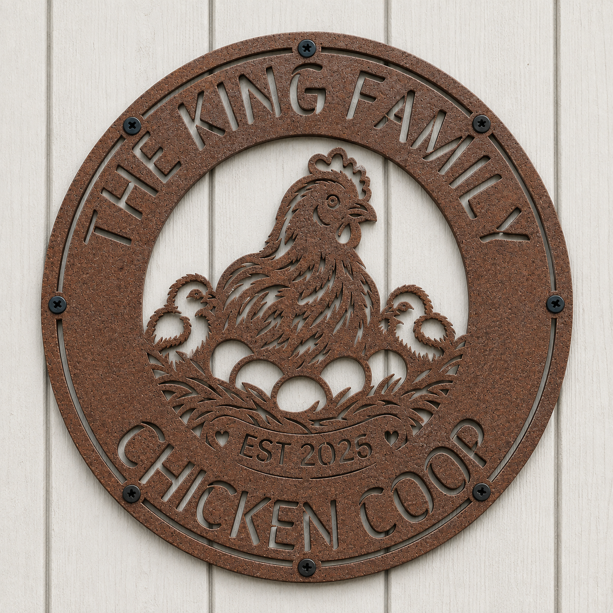 A round metal chicken coop plaque with a textured brown finish, featuring a laser-cut design of a hen with eggs and the text 'THE KING FAMILY CHICKEN COOP EST 2025', mounted on a white wood paneled wall.
