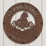 A round metal chicken coop plaque with a textured brown finish, featuring a laser-cut design of a hen with eggs and the text 'THE KING FAMILY CHICKEN COOP EST 2025', mounted on a white wood paneled wall.