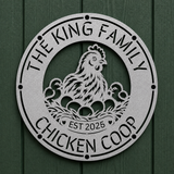 A round metal garden sign featuring a chicken and two chicks with eggs in a nest, surrounded by the text 'THE KING FAMILY CHICKEN COOP' and 'EST 2025'. The sign is mounted on a dark green wood-paneled wall.