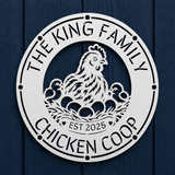 A round white metal sign featuring a cutout illustration of a hen, two chicks, and eggs, with the text 'THE KING FAMILY CHICKEN COOP' and 'EST 2025', mounted on a dark blue wood plank background.