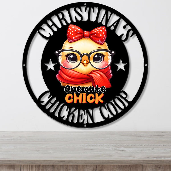 A circular black metal wall sign featuring a cartoon chick wearing a red polka-dot bow and red scarf, with the text 'CHRISTMAS CHICKEN COOP' and 'one cute CHICK' in white and orange lettering, shown hanging on a light gray wall above a wooden surface.