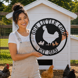 A woman in a white t-shirt and beige overalls stands in front of a white chicken coop, holding up a circular black metal sign that reads 'OUR ANIMALS FOOD EST 2020' with a silhouette of a chicken and chicks.