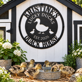 A decorative black metal sign reading 'Kristine's Lucky Ducks Quack House Est. 2026' mounted on a white and black chicken coop, with several ducklings gathered around a galvanized metal water trough in front.