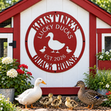 A decorative red and white metal sign reading 'Lucky Ducks Quack House' mounted on a small white and red shed, with a white duck, three ducklings, and a brown mallard duck standing in front of it outdoors.