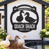 A black metal decorative sign reading 'AMANDA'S QUACK SHACK SINCE 2025' with duck silhouettes, mounted on a white shed. Three live ducks stand in front of the shed near a small pond, with flowers on both sides.