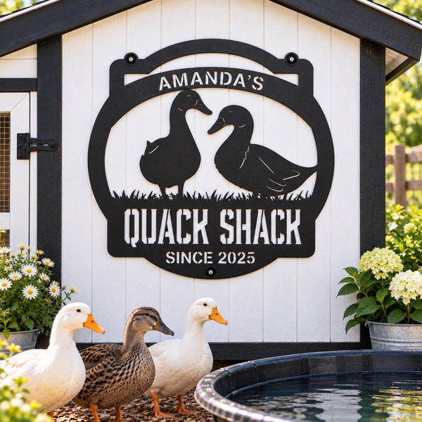 A black metal decorative sign reading 'AMANDA'S QUACK SHACK SINCE 2025' with duck silhouettes, mounted on a white shed. Three live ducks stand in front of the shed near a small pond, with flowers on both sides.