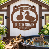 A rustic metal sign reading 'Amanda's Quack Shack Since 2025' with duck silhouettes, mounted on a white and brown chicken coop. Ducks, including several ducklings, swim in a small pond in front of the coop.