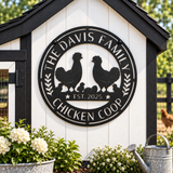 A circular black metal sign mounted on a white chicken coop wall, featuring silhouettes of two chickens, a chick, wheat ears, and the text 'THE DAVIS FAMILY CHICKEN COOP' and 'EST. 2025'.