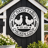 A circular black metal sign mounted on a black wooden chicken coop door, featuring white silhouettes of two chickens, several chicks, and the text 'THE DAVIS FAMILY CHICKEN COOP' and 'EST. 2025'.
