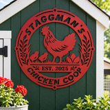 A red metal decorative sign shaped like a chicken coop, mounted on a dark green wooden shed wall. The sign features a silhouette of a rooster and two chicks with eggs, the text 'STAGGMAN'S CHICKEN COOP', and 'EST.