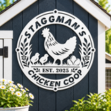 A large white circular metal sign featuring a chicken and two chicks, flowers, and the text 'STAGGMAN'S CHICKEN COOP' and 'EST. 2025', mounted on the front of a dark blue shed with white trim and green plants in the foreground.