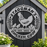 A large circular galvanized metal sign mounted on a dark gray wooden chicken coop, featuring a rooster, two chicks, eggs, and the text 'STAGGMAN'S CHICKEN COOP' and 'EST. 2025'.